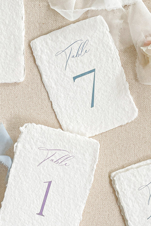 Bring elegance to your reception with our folded handmade paper table numbers. Crafted from luxurious deckled-edge cotton paper, each card features modern calligraphy paired with soft, timeless colors to guide guests effortlessly. Their folded construction allows them to stand on their own, creating a refined and cohesive tablescape for weddings, celebrations, and fine-art events.