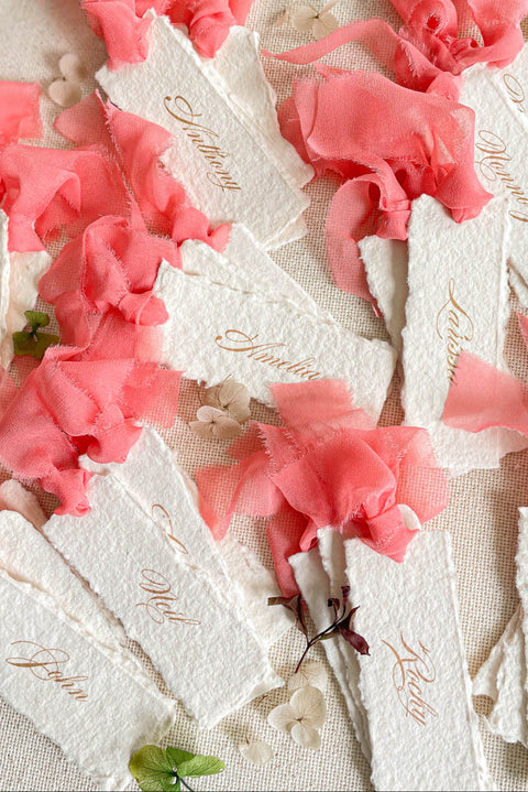 Bring a lively, romantic touch to your event with our handmade paper place cards, featuring natural deckled edges, vibrant coral chiffon ribbon, and elegant gold calligraphy. Crafted individually for a fine-art, textural finish, these name cards elevate any wedding tablescape with their vivid color and artisanal charm. Perfect for modern, garden, summer, and destination weddings.