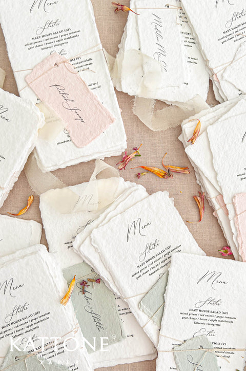Overhead close-up of elegant ivory-white handmade paper wedding menus featuring textured deckled edges, black calligraphy, and classic serif typography. The menus are paired with personalized handmade paper place cards, secured with delicate natural twine. Soft blush pink and sage green accents, ivory chiffon ribbon, and scattered flower petals complete the romantic wedding stationery arrangement on a beige linen background.