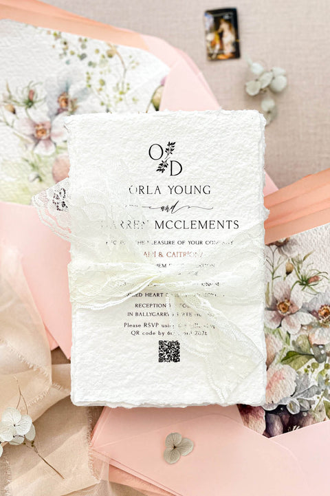 Luxury handmade cotton paper wedding invitation with deckled edges wrapped in ivory lace ribbon, styled with blush pink envelopes and watercolor floral envelope liners.