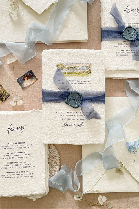 Luxury handmade paper wedding invitation suite featuring deckled edge cotton paper, custom watercolor venue artwork, dusty blue chiffon ribbon wraps, monogram wax seals, itinerary cards, and coordinated ivory envelopes.