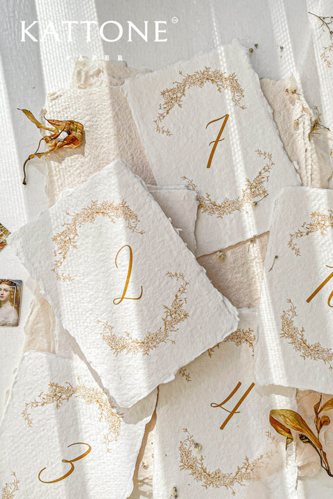 These handmade cotton paper table numbers feature elegant gold numbering framed by intricate botanical artwork, creating a warm and refined fine-art aesthetic. The soft deckle edges and textured surface add authentic artisanal character, making them perfect for romantic, nature-inspired, or luxury wedding themes. Designed to enhance your tables with organic beauty and timeless sophistication, they offer both practicality and artistry.