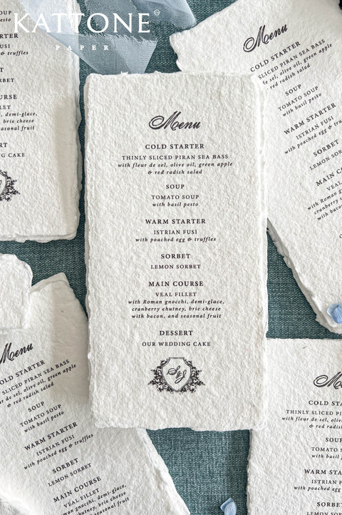 Close-up overhead photograph of a luxury ivory-white handmade paper wedding menu featuring textured deckled edges, an elegant black calligraphy heading, and classic serif typography. The menu displays multiple wedding dinner courses and a personalized floral shield monogram at the bottom. Coordinating menus are arranged around it on a muted blue textured background with soft blue chiffon ribbon and delicate blue flower petals.