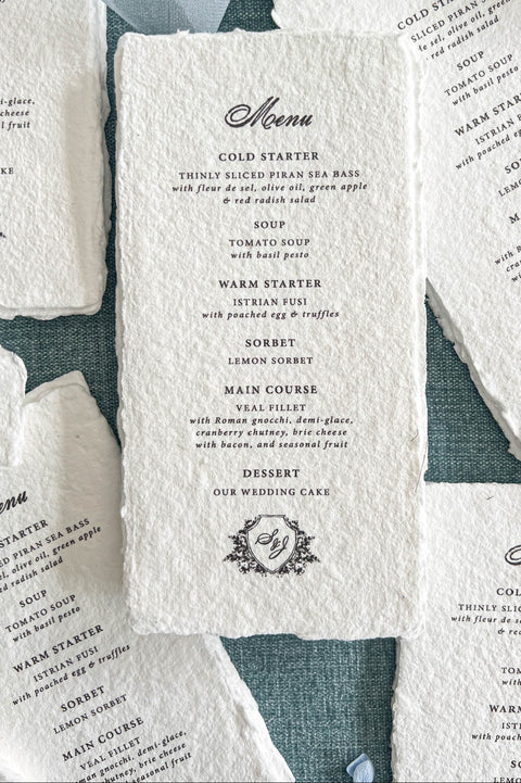 Close-up overhead photograph of a luxury ivory-white handmade paper wedding menu featuring textured deckled edges, an elegant black calligraphy heading, and classic serif typography. The menu displays multiple wedding dinner courses and a personalized floral shield monogram at the bottom. Coordinating menus are arranged around it on a muted blue textured background with soft blue chiffon ribbon and delicate blue flower petals.