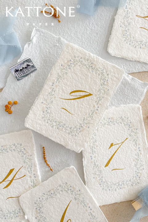 Hand-painted floral wreaths and artisanal deckle-edge cotton paper come together to create table numbers with a soft, fine-art aesthetic. Each card features delicate watercolor detailing paired with elegant gold numerals, offering a timeless blend of natural beauty and refined craftsmanship. Perfect for garden weddings, spring celebrations, or romantic fine-art events, these table numbers bring an ethereal charm to every tablescape.