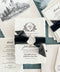 Overhead flat lay of a luxury black and white wedding invitation suite featuring ivory handmade paper invitations with personalized floral crest monograms, black sheer ribbons, and embossed ivory wax seals. Coordinating wedding details cards, ivory envelopes with black architectural illustrations of a historic lakeside church, elegant address printing, and soft blue ribbon accents are arranged on a textured blue-green background.