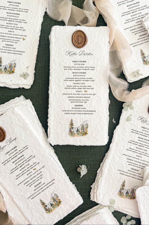Overhead arrangement of personalized handmade wedding dinner menus with naturally deckled edges, individual guest names, antique gold monogram wax seals, and delicate watercolor pine tree illustrations, displayed with flowing ivory chiffon ribbon and scattered pale green flowers on a dark green background.