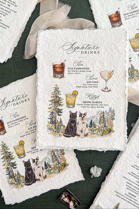 Overhead flat lay of personalized handmade paper wedding signature cocktail menus featuring illustrated drinks, a black cat and a white cat with dark markings, and watercolor forest scenery. The deckled-edge menus are arranged alongside matching wedding dinner menus, flowing ivory ribbon, and delicate flowers on a forest green background.