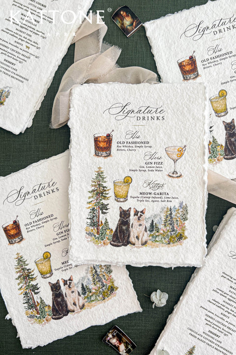 Overhead flat lay of personalized handmade paper wedding signature cocktail menus featuring illustrated drinks, a black cat and a white cat with dark markings, and watercolor forest scenery. The deckled-edge menus are arranged alongside matching wedding dinner menus, flowing ivory ribbon, and delicate flowers on a forest green background.