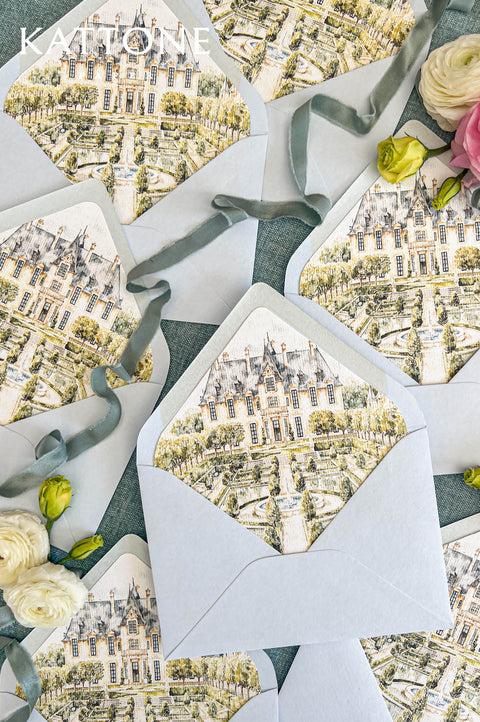 Powder blue wedding envelopes with custom watercolor château liners featuring an elegant European manor, formal gardens, ornamental fountain, and landscaped terraces. Close-up flat lay with dusty blue silk ribbon and cream flowers on a textured blue background.