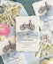 Flat lay of luxury Italian destination wedding save the date cards printed on textured ivory handmade paper with natural deckled edges and detailed black architectural castle illustrations. The cards are tied with dusty blue chiffon ribbon and accompanied by pale blue envelopes with colorful watercolor château and garden liners, surrounded by romantic flowers and decorative ribbon.