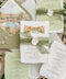 Overhead flat lay of a luxury sage green wedding invitation suite featuring white deckled handmade paper, watercolor tropical resort illustration, olive green envelopes with illustrated liners, matching weekend details cards, silk ribbon and ivory monogram wax seals.