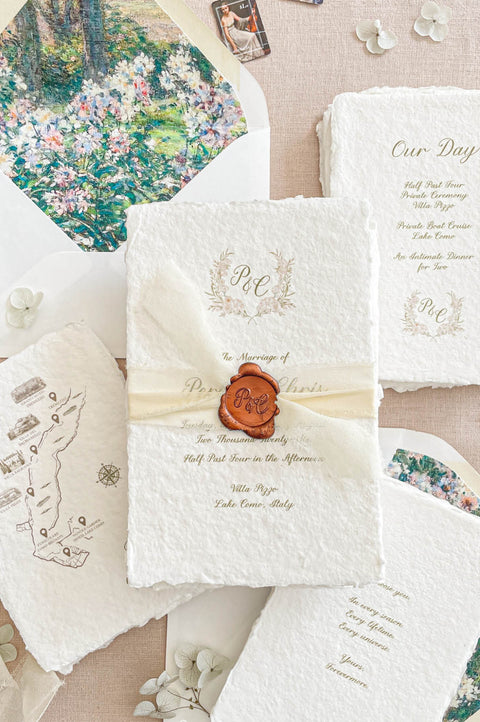 A styled flat-lay of Kattone Papers wedding stationery, featuring a central ivory textured invitation sealed with a brown wax seal, surrounded by coordinating cards, an envelope with floral artwork, and a map card on a beige fabric background.