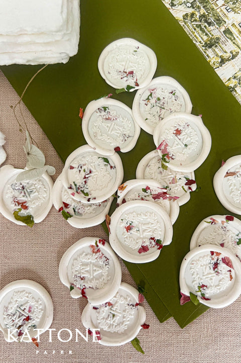 Close-up top-down view of ivory floral wedding wax seals with embossed monogram initials and embedded pink, red, and green dried flower petals, arranged on olive green paper and neutral linen