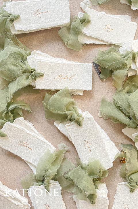 Top-down flat lay of personalized handmade paper wedding place cards with textured deckled edges, warm calligraphy guest names, and long sage green frayed silk ribbon ties, styled with dried flowers on a neutral linen background