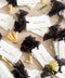 Top-down flat lay of personalized handmade paper wedding place cards with deckled edges, each printed with guest names in soft brown calligraphy, styled across neutral linen with sheer black ribbon and scattered yellow flowers for an elegant reception table setting display