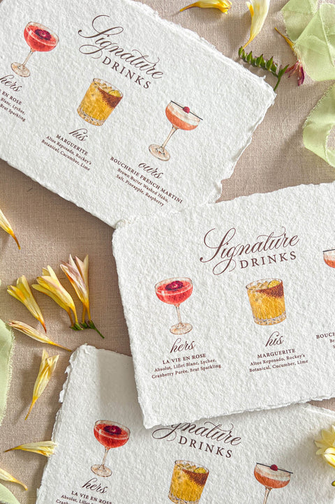 Top-down flat lay of handmade deckled-edge signature drinks wedding menus displayed on a neutral linen background, featuring watercolor cocktail illustrations, elegant burgundy script lettering, soft serif text, scattered yellow flowers, and pale green silk ribbon, styled as romantic custom wedding bar menu stationery