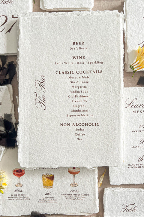 Top-down flat lay of handmade deckled-edge wedding bar menu printed on textured ivory paper, featuring burgundy typography with beer, wine, classic cocktails, and non-alcoholic drink selections, surrounded by table numbers, signature cocktail cards, black ribbon, and yellow floral styling