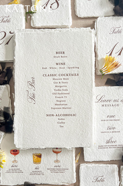 Top-down flat lay of handmade deckled-edge wedding bar menu printed on textured ivory paper, featuring burgundy typography with beer, wine, classic cocktails, and non-alcoholic drink selections, surrounded by table numbers, signature cocktail cards, black ribbon, and yellow floral styling