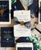 A styled flat-lay of wedding stationery from Kattonal, featuring navy blue and cream textured paper invitations, envelopes, a wax seal, botanical prints, and small photo cards arranged on a neutral background.