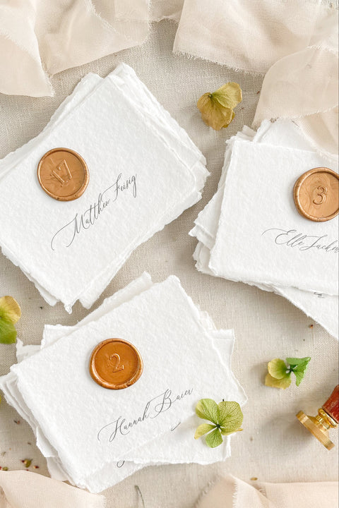 Add an artisanal charm to your wedding tables with our handmade paper place cards, delicately crafted from 100% cotton paper with natural deckled edges. Finished with elegant calligraphy and a wax seal detail, these fine-art cards bring a sense of timeless romance and sophistication. Perfect for luxury weddings, garden receptions, or intimate dinner parties, they seamlessly combine old-world craftsmanship with modern elegance.