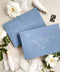 Dusty blue wedding envelopes with printed addresses, styled flat lay with white florals, ribbon, and greenery, perfect for elegant and coastal invitation suites.