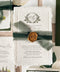 Wedding stationery arranged in a luxury flat lay showcasing handmade paper invitations, vellum wraps, green backing cards, ribbon, wax seals, and coordinated inserts.