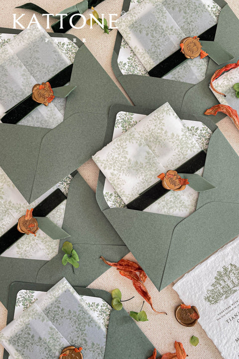 Flat lay of soft green envelopes with watercolor floral liners styled with ribbon, greenery, and fine art wedding stationery details.