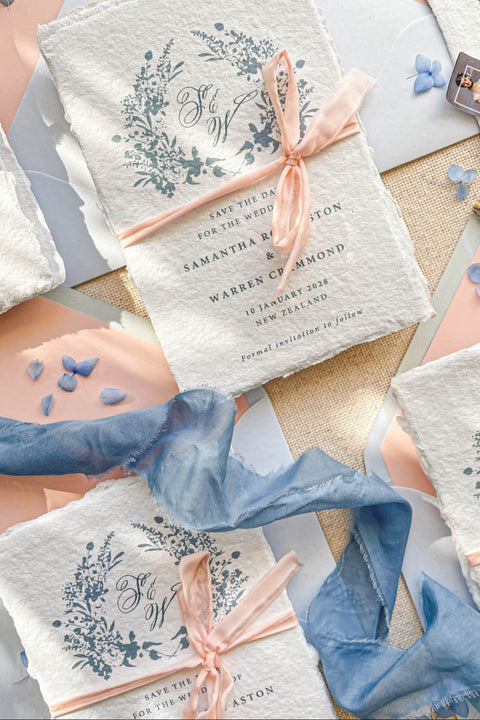 Announce your wedding with elegance and artistry through our handmade paper Save the Date cards, adorned with a soft blue watercolor floral wreath and tied with a delicate blush silk ribbon. Each card is crafted on luxury 100% cotton handmade paper with naturally torn deckled edges, creating a timeless and romantic keepsake that beautifully reflects your love story.