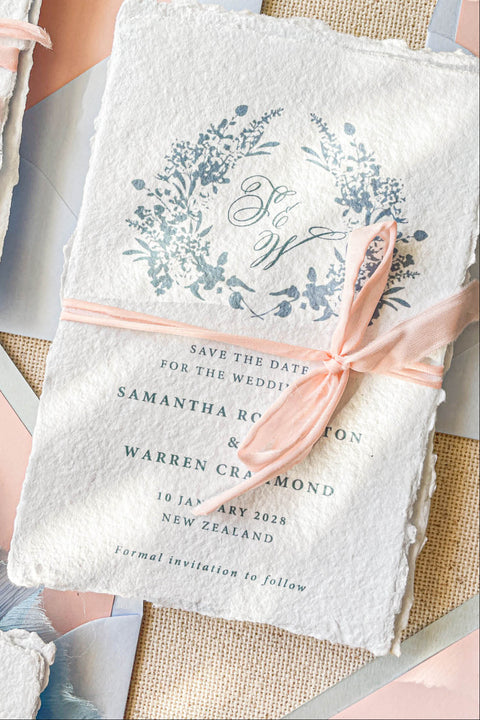 Announce your wedding with elegance and artistry through our handmade paper Save the Date cards, adorned with a soft blue watercolor floral wreath and tied with a delicate blush silk ribbon. Each card is crafted on luxury 100% cotton handmade paper with naturally torn deckled edges, creating a timeless and romantic keepsake that beautifully reflects your love story.