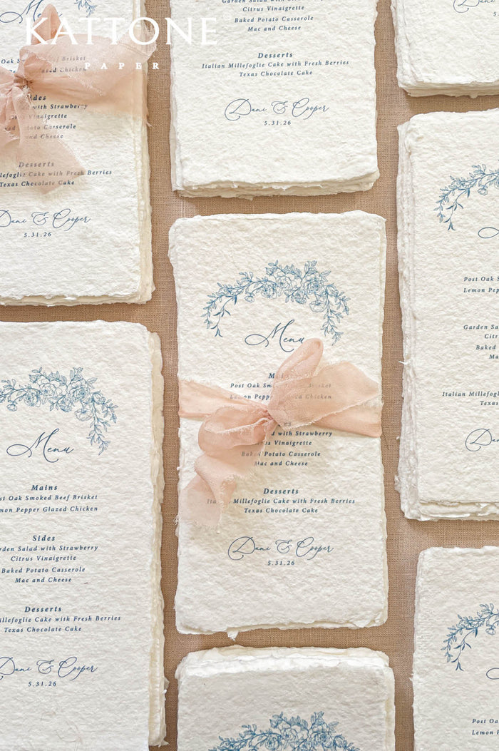 Luxury handmade paper wedding menus featuring deckled edge cotton paper, blue floral artwork, personalized guest names, and blush chiffon ribbon styling.