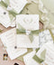 Luxury handmade cotton paper wedding invitation with deckled edges wrapped in sage silk ribbon and wax seal, styled with botanical envelope liners and soft green envelopes.