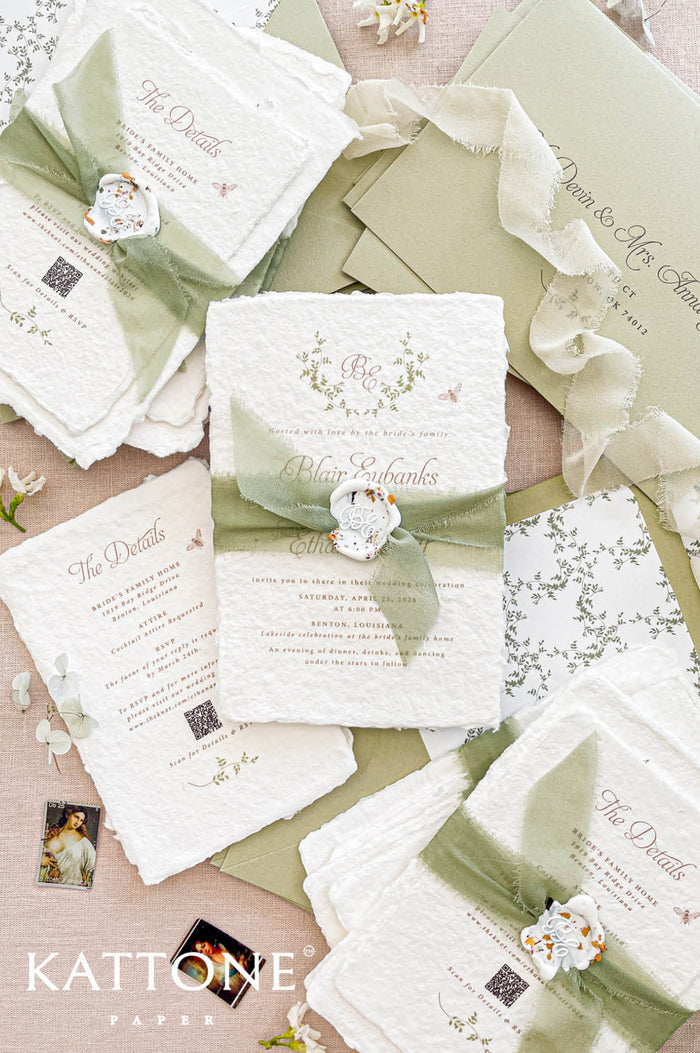 Luxury handmade cotton paper wedding invitation with deckled edges wrapped in sage silk ribbon and wax seal, styled with botanical envelope liners and soft green envelopes.