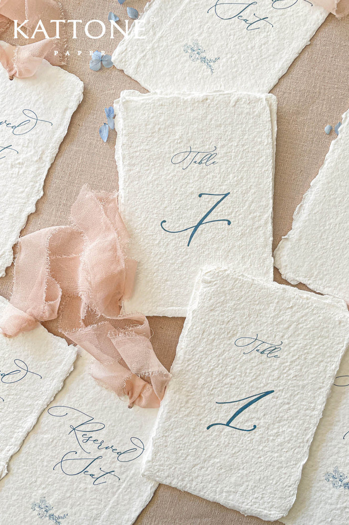 Luxury handmade paper reserved seat cards and table numbers featuring deckled edge cotton paper, elegant blue typography, and blush chiffon ribbon styling.