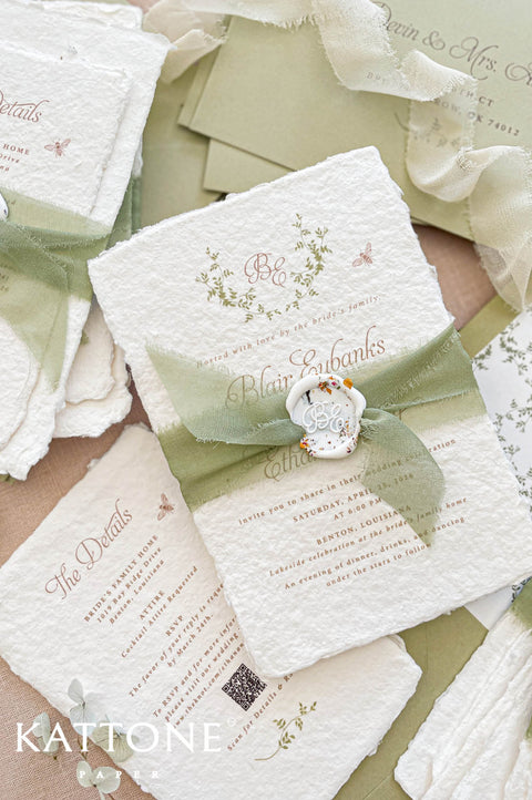Close up of textured handmade cotton paper wedding invitation featuring botanical monogram wreath and elegant serif typography.