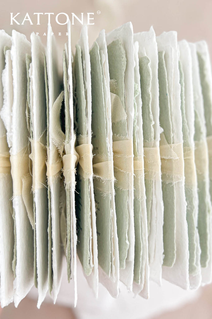 Side view of a collection of luxury handmade paper wedding invitations and matching sage green details cards, showcasing thick textured paper, naturally irregular deckled edges, alternating ivory and pale green layers, and delicate cream ribbon wrapping.