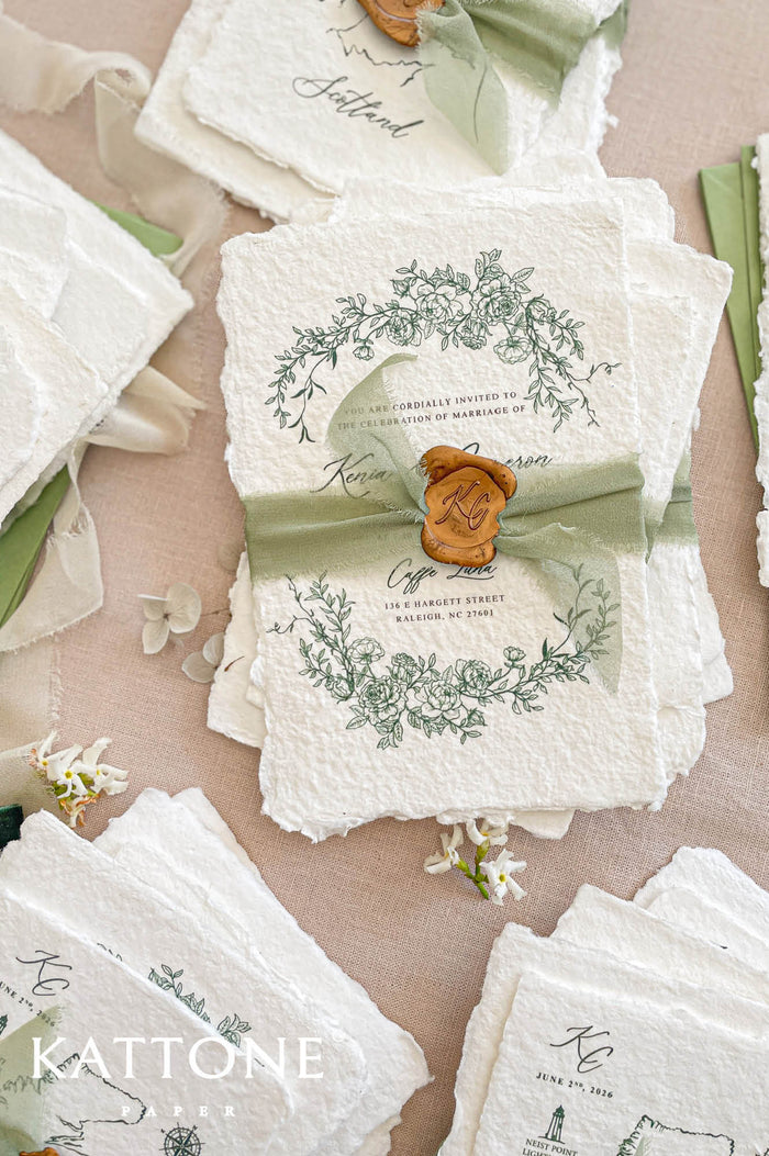 Luxury handmade cotton paper wedding invitation with deckled edges and botanical wreath, styled with sage silk ribbon, wax seal and coordinated stationery suite on neutral linen background.