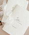 Close-up photograph of personalized ivory handmade paper wedding programs with naturally deckled edges, elegant black calligraphy reading To Allison on our Wedding Day, and a printed October wedding date. The minimalist wedding stationery is tied with flowing sheer ivory ribbon bows and arranged on a soft beige linen background.