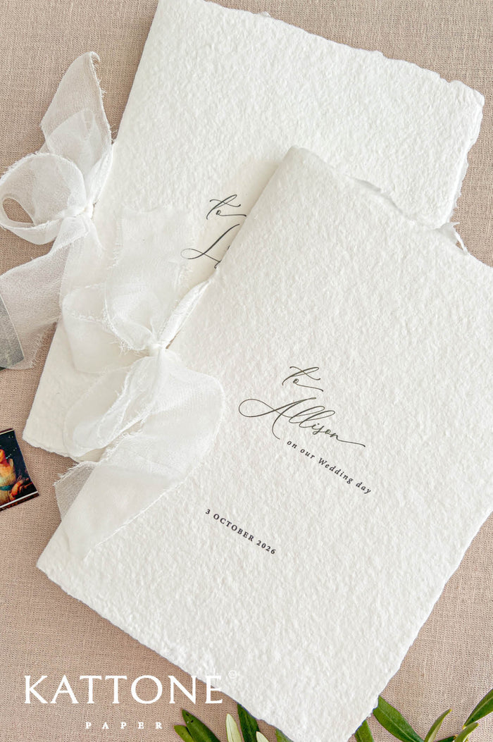 Close-up photograph of personalized ivory handmade paper wedding programs with naturally deckled edges, elegant black calligraphy reading To Allison on our Wedding Day, and a printed October wedding date. The minimalist wedding stationery is tied with flowing sheer ivory ribbon bows and arranged on a soft beige linen background.