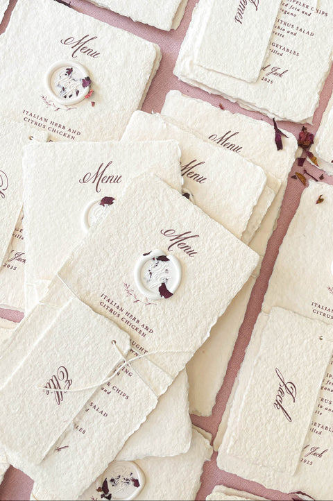 Stacked handmade paper wedding menus with deckled edges, wax seal accents, and personalized name tags.