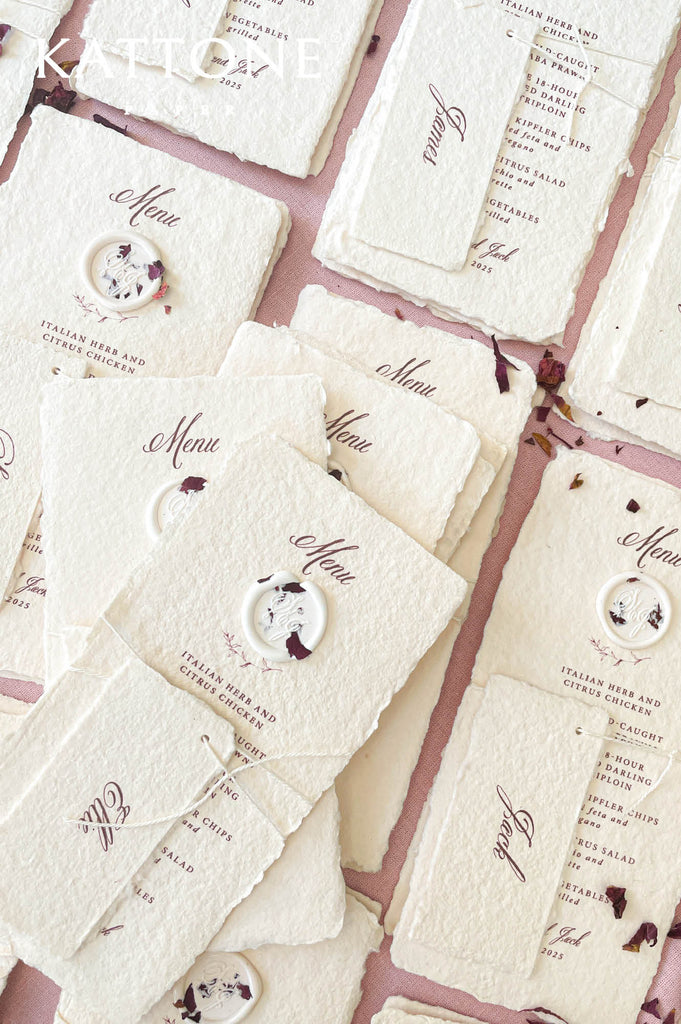 Stacked handmade paper wedding menus with deckled edges, wax seal accents, and personalized name tags.