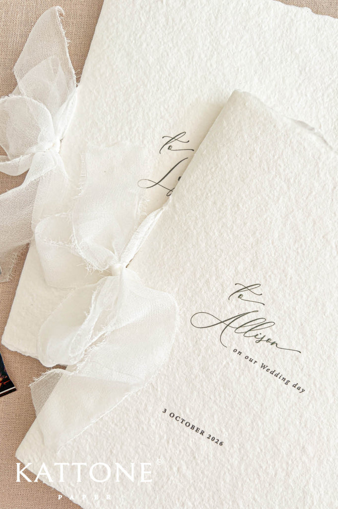 Overhead flat lay of elegant handmade paper wedding ceremony programs featuring thick textured ivory paper, organic deckled edges, personalized calligraphy, and delicate sheer ivory ribbon bows. Two overlapping minimalist wedding programs are displayed on natural beige fabric with subtle green foliage along the bottom edge.
