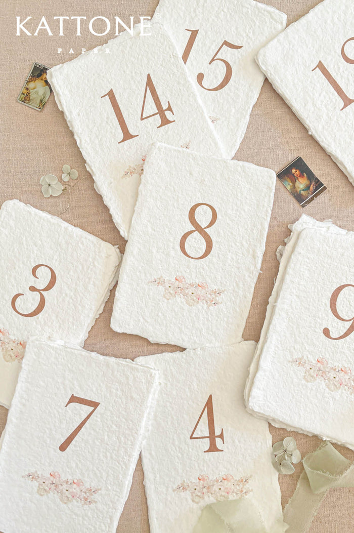Handmade wedding table numbers on deckled edge cotton paper with warm terracotta numbering and soft floral detail in fine art wedding style.