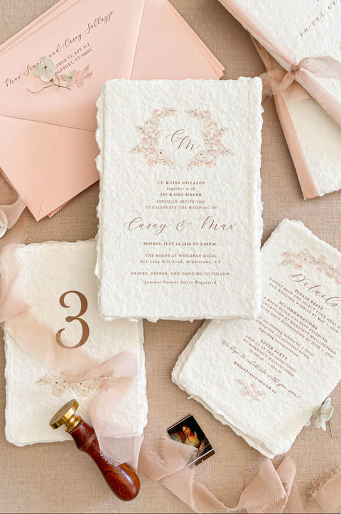 Luxury handmade cotton paper wedding invitation with deckled edges and blush floral wreath, styled with silk ribbon, wax seal, blush envelopes and coordinated stationery suite.