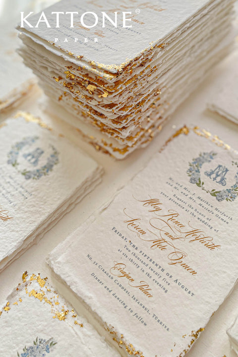 This handmade paper wedding invitation suite combines fine craftsmanship with opulent detailing. Printed on luxury cotton paper with deckled edges, each invitation is accented with subtle touches of gold leaf, catching the light with a refined shimmer that speaks of romance and sophistication. The design features a hand-painted floral monogram, symbolizing the couple’s union, surrounded by soft watercolor greenery and pastel hues.