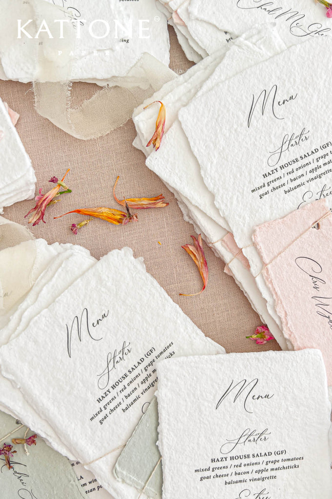 Overhead close-up of elegant ivory-white handmade paper wedding menus featuring textured deckled edges, black calligraphy, and classic serif typography. The menus are paired with personalized handmade paper place cards, secured with delicate natural twine. Soft blush pink and sage green accents, ivory chiffon ribbon, and scattered flower petals complete the romantic wedding stationery arrangement on a beige linen background.