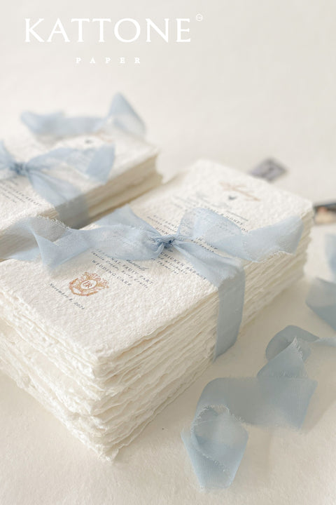 Refined, romantic, and artisanal - our handmade paper wedding menus bring a touch of timeless elegance to your table setting. Each menu is printed on 100% cotton paper with naturally torn deckled edges, offering a beautifully textured surface that speaks of craftsmanship and care. Tied with a soft dusty blue silk ribbon and adorned with a subtle crest monogram, these menus perfectly complement fine art, coastal, and destination weddings.