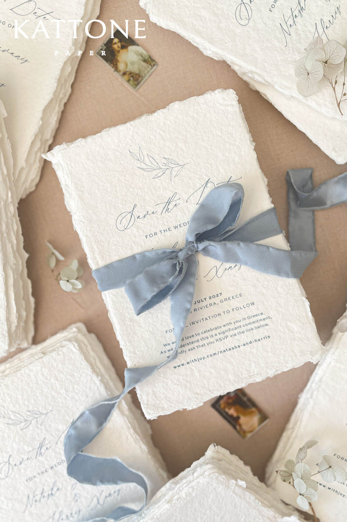 Dusty blue silk ribbon tied around handmade save the date card creating a soft romantic bow on textured deckled edge paper.