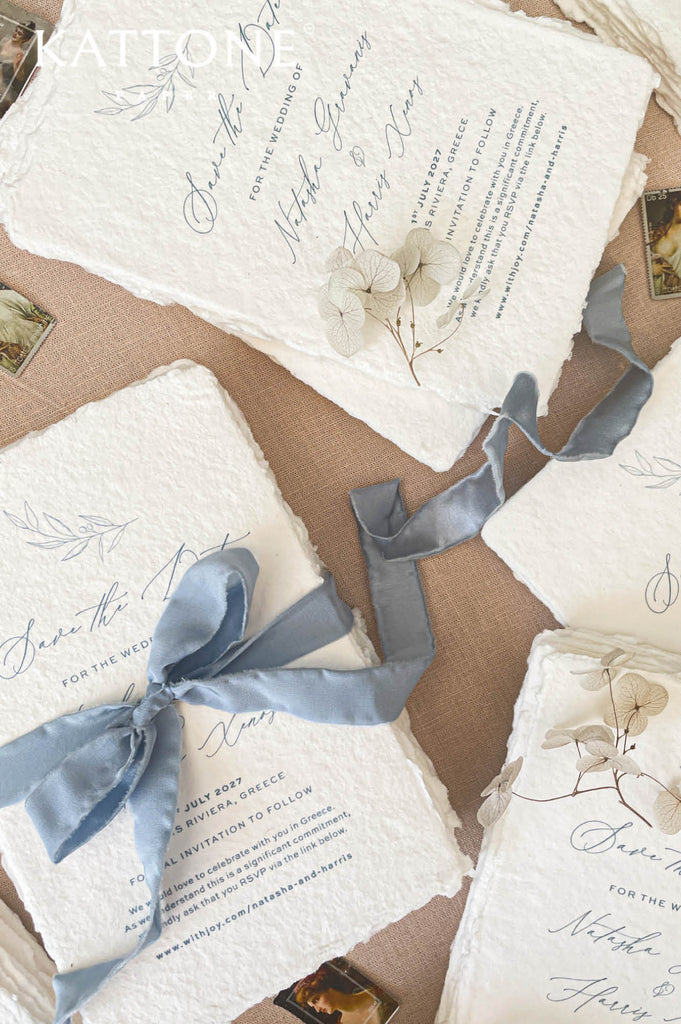 Dusty blue silk ribbon tied around handmade save the date card creating a soft romantic bow on textured deckled edge paper.