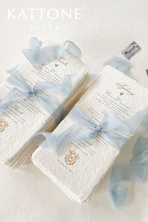 Refined, romantic, and artisanal - our handmade paper wedding menus bring a touch of timeless elegance to your table setting. Each menu is printed on 100% cotton paper with naturally torn deckled edges, offering a beautifully textured surface that speaks of craftsmanship and care. Tied with a soft dusty blue silk ribbon and adorned with a subtle crest monogram, these menus perfectly complement fine art, coastal, and destination weddings.